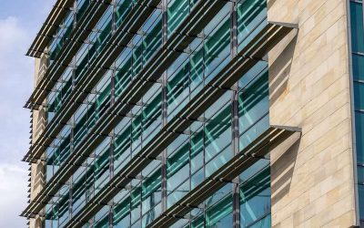 Modern Building Facade with Stone and Glass Windows  in Newcastle upon Tyne, UK. A modern building facade features tan stone and reflective blue-green glass windows with horizontal metal shades, under a cloudy sky  in Newcastle upon Tyne, UK.