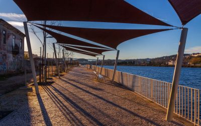 A paved walkway with shade sails runs alongside a river, offering a scenic view of the water and distant hills