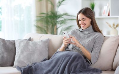 Happy woman in winter at home using phone covered by blanket