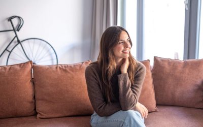 A smiling young woman sits on a plush couch in a cozy, modern living room, looking out the window as soft natural light brightens the space and creates a relaxed, content atmosphere.