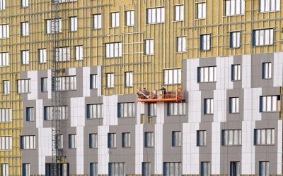 Workers team on construction cradle cladding and warming wall of monolithic house under construction. Man on suspended platform insulating wall facade of residential building.